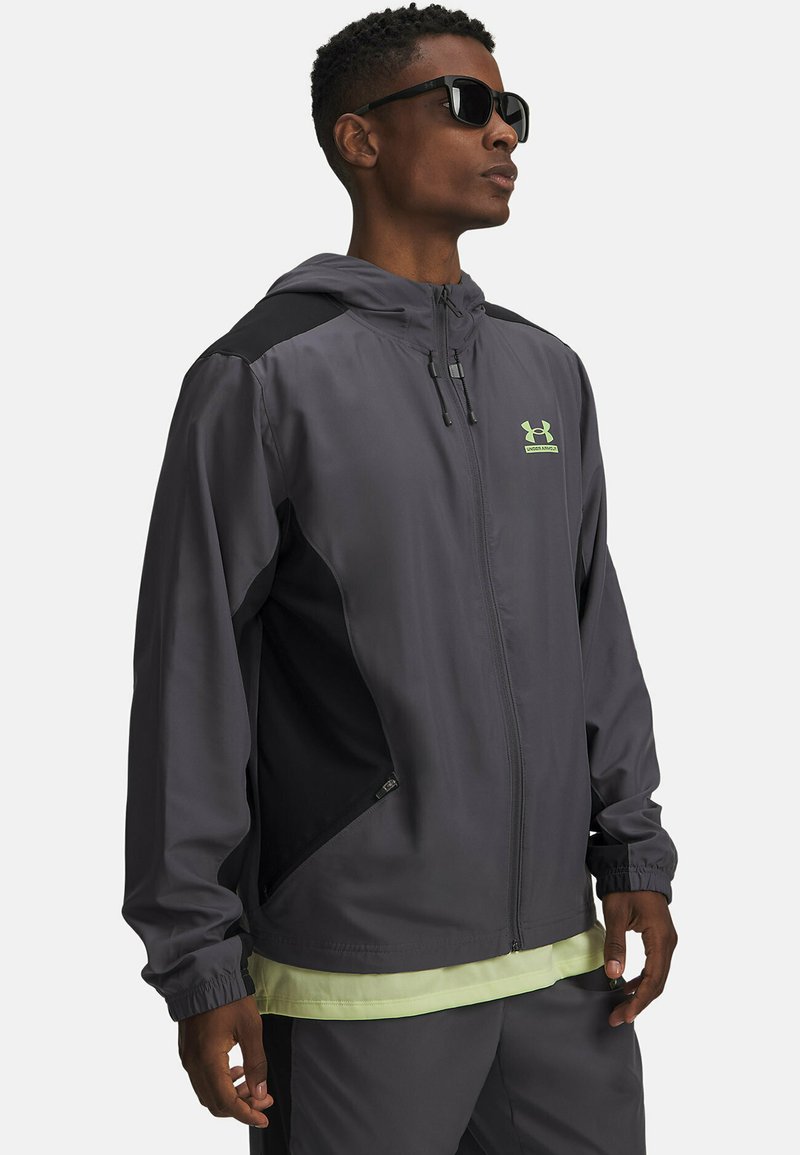 Grijze capuchonjas met zwarte accenten, voorzien van een rits aan de voorkant, elastische manchetten en een reflecterend logo op de borst. Lichtgewicht stof.