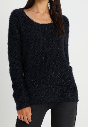 Femme portant un pull en maille noir douillet à manches longues et à encolure ronde, associé à un pantalon noir.