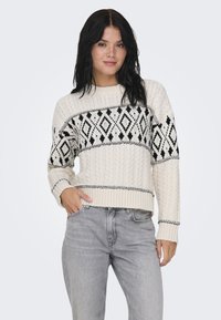 Kremfarget strikkegenser med svart geometrisk mønster, ribbede mansjetter og hem; kombinert med lysegrå jeans. Myk tekstur, avslappet passform.