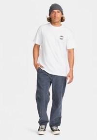 Camiseta branca em algodão com decote redondo e estampa de logotipo no peito, combinada com calças de veludo cotelê azul-escuras e sapatilhas preto e brancas.