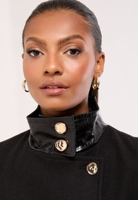 Sort frakke med høj krave med krokodilleteksturerede detaljer, guldknapper og en glat stoffinish.