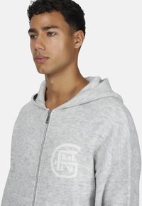 Szary sweter z kapturem zapinany na zamek wykonany z miękkiego materiału, z dopasowanym kapturem, zamkiem z przodu oraz logo na lewej piersi.