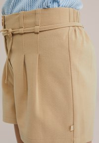 Beiga shorts i mjukt tyg har en veckad design, justerbart snöre i midjan och en liten broderad logotyp på sidan.