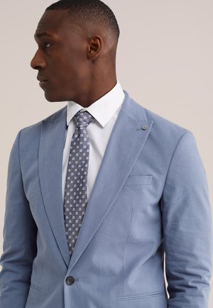 Bărbat în sacou bleu deschis, cămașă albă și cravată cu model, privind spre stânga pe un fundal simplu.