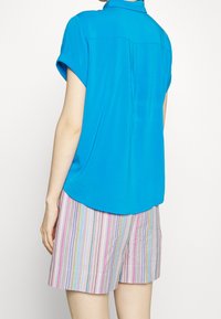 Maglietta azzurra a manica corta con orlo arrotondato, abbinata a pantaloni corti a righe colorate con linee verticali rosa, blu e bianche.