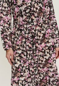 Bloemmotief jurk in zwart, roze en crème. Kenmerken zijn lange mouwen, een ceintuur in de taille en geplooide details in de rok. Geribbelde stof.