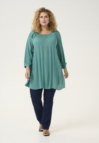 Blusa leggera color turchese con scollo rotondo, dettaglio a chiave e maniche lunghe, abbinata a jeans blu scuro e sandali neri.