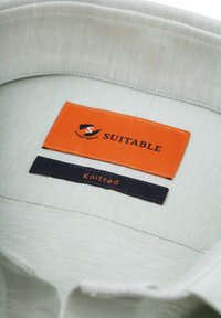 Gehaakte kraaglabel in lichtgroene stof, met een oranje "Geschikt" label en een zwart "Gehaakt" label met oranje letters eronder.