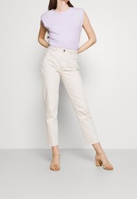 Woman wearing a sleeveless lilac top, high-waisted light beige jeans, and beige block-heeled sandals, standing with hands on hips.