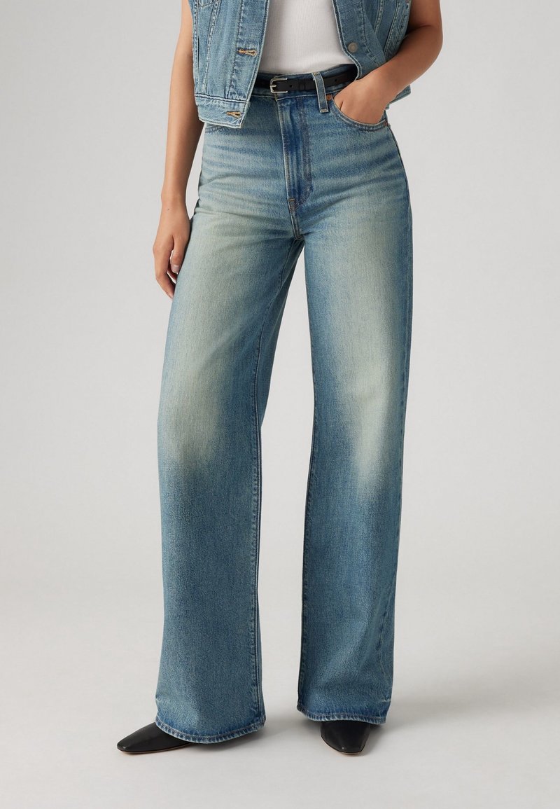 Jean large taille haute bleu clair avec une finition délavée et des coutures contrastantes, associé à une ceinture noire et des chaussures pointues noires.