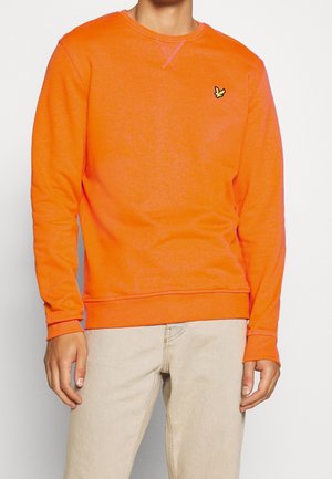 Person wearing a bright orange long-sleeve sweatshirt with a small yellow bird logo on the left chest and beige pants.