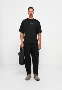 Man in zwarte "Studio Edition" T-shirt en zwarte cargo-broek, met een zwarte jas in de hand, staande tegen een witte achtergrond.