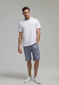 Biała koszulka polo z wzorzystym kołnierzykiem i logo na piersi, zestawiona z jasnoniebieskimi szortami do kolan oraz białymi trampkami.