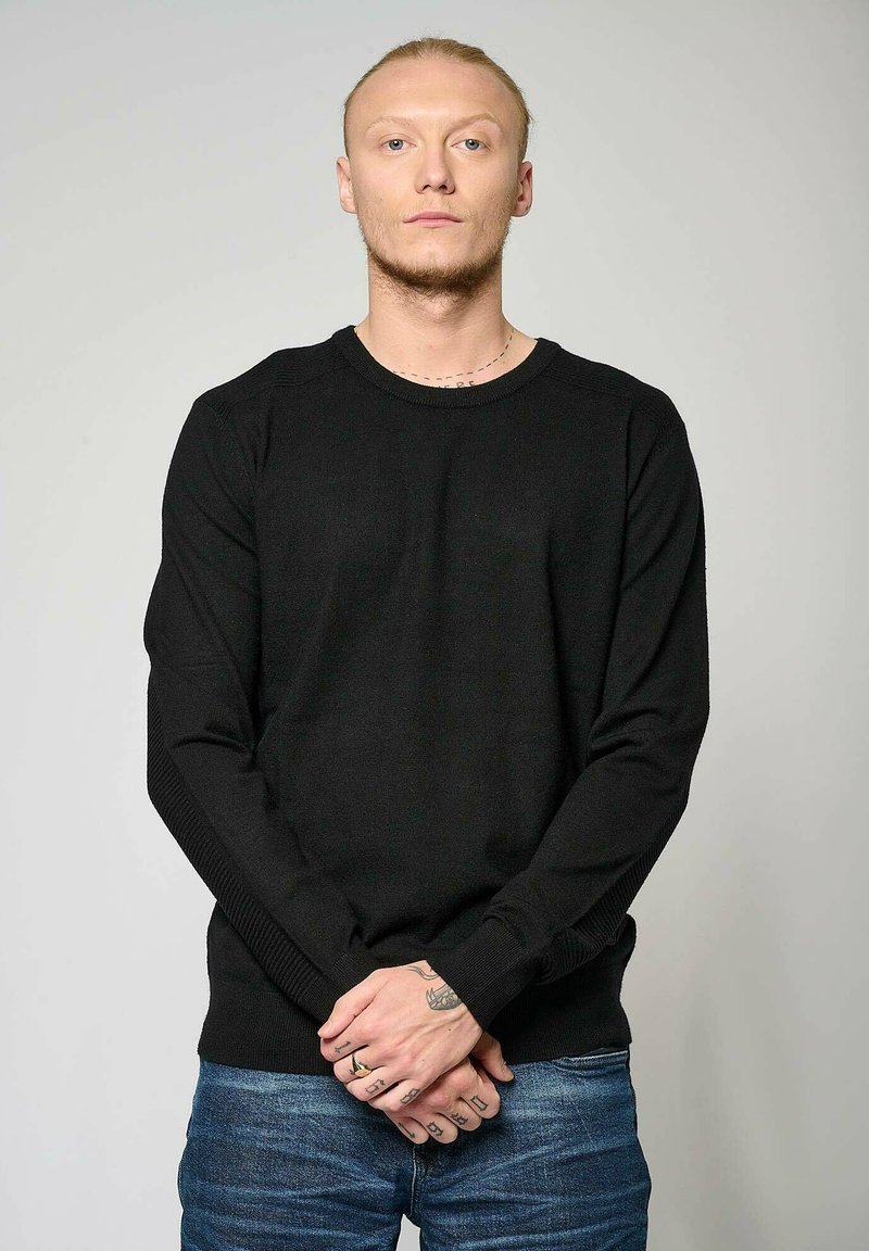 Pull noir à manches longues avec un col rond côtelé et des panneaux latéraux texturés, porté avec un jean bleu.