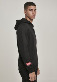 Haut à capuche noir avec une poche avant, orné d'un logo NASA rouge sur la manche. Fabriqué en tissu doux, avec un cordon de serrage et des poignets côtelés.