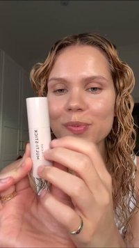 Baume à lèvres dans un tube rond et blanc avec un texte noir ; texture lisse et finition brillante ; tenu par une personne avec des cheveux bouclés et mouillés.