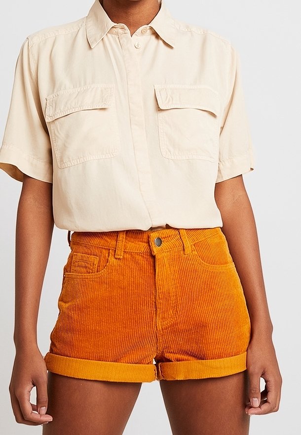 Personne portant une chemise beige à manches courtes boutonnée rentrée dans un short en velours côtelé orange taille haute avec des revers roulés.