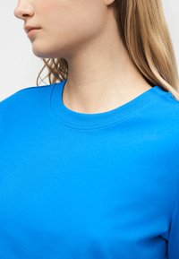 Giovane con capelli lunghi biondi che indossa una maglietta a girocollo di un brillante blu, visibile dal collo alla parte superiore del petto.