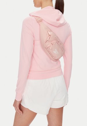 Kobieta w różowej bluzie z kapturem i białych szortach, z jasnoróżową saszetką Reebok przewieszoną przez plecy, na białym tle.