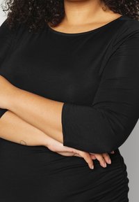 Haut noir à manches trois-quarts en matière douce et extensible. Doté d'un col rond et d'un détail froncé sur le côté pour une coupe ajustée.