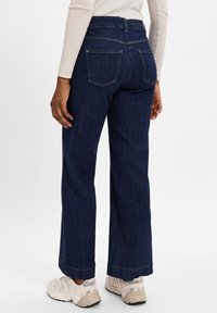 Mørkeblå denim bukser med vid benvidde og middels høyde midje, med baklommer og cropped nederkant, kombinert med beige joggesko.