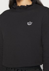 Svart cropped långärmad tröja med volangkrage. Har en vit broderad logotyp på bröstet. Mjuk bomullsmaterial.