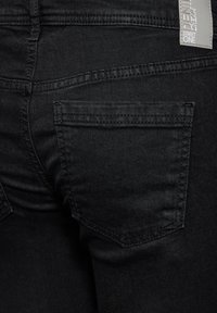 Svarta denimjeans med en närbild av bakfickan, med synlig sömnad och en etikett, som visar en slät textur och en åtsittande design.