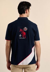 Polo de color navy con un diseño curvado en rojo y blanco, que presenta un gran logo bordado rojo en la espalda, realzado con un escudo y texto.