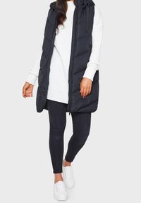 Zwarte gewatteerde bodywarmer met capuchon, doorgestikte textuur, rits aan de voorkant, gecombineerd met een witte gebreide trui en zwarte legging, aangevuld met witte sneakers.