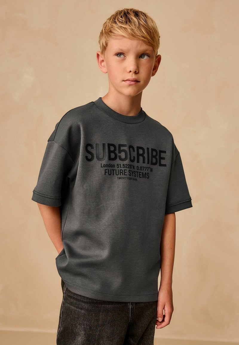 T-shirt grigio oversize a maniche corte con testo grafico nero, che presenta le coordinate e la frase "FUTURE SYSTEMS". Tessuto morbido; design casual.
