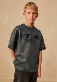 T-shirt grigio oversize a maniche corte con testo grafico nero, che presenta le coordinate e la frase "FUTURE SYSTEMS". Tessuto morbido; design casual.