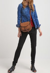 Hnědá kožená crossbody taška s klopou, detail se třásněmi a prošívanými akcenty. V kombinaci s denimovou bundou, červeným topem a černými kalhotami.