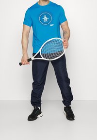 Man i blå T-shirt och marinblå joggingsbyxor som håller en vit tennisracket, stående mot en enkel ljus bakgrund.