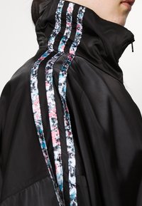 Svart vindjacka med hög krage, tre färgglada randiga detaljer i rosa och blått på axeln, mjuk textur, dragkedjestängning.