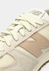 Gros plan sur une basket New Balance beige avec des lacets crème, un tissu en mesh, des détails en daim et un logo "N" cousu sur le côté.
