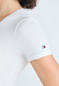 Bílý krátký rukáv tričko vyrobené z hladké látky, s malým vyšitým logem červené, bílé a modré barvy na levém rukávu.