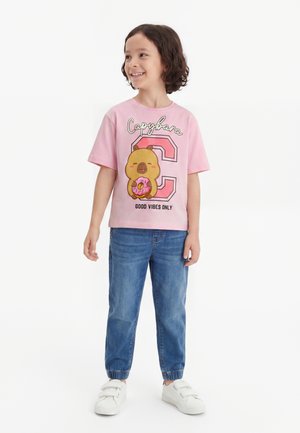 Dziecko z kręconymi włosami, ubrane w różowy t-shirt z kapibarą trzymającą pączka, niebieskie dżinsy i białe trampki, uśmiechające się i patrzące na bok.
