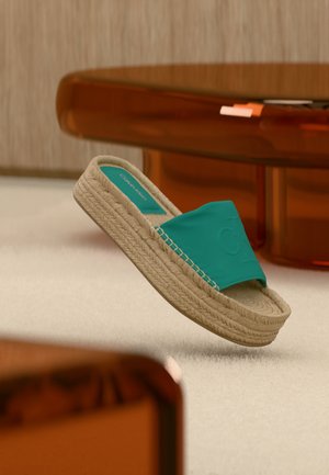 Zelenomodrá slide sandála s tkanou béžovou platformovou podrážkou ležiaca na svetlom koberci, blízko dreveného stola s oranžovými akcentmi.