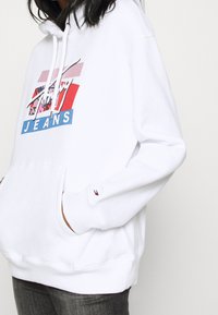 Biała bluza z kapturem z kolorowym wzorem graficznym z napisem "Jeans". Posiada kieszeń na przodzie oraz regulowany kaptur. Wyposażona w małe logo.