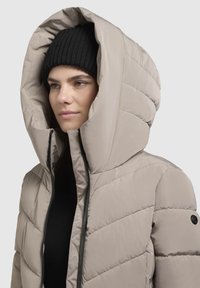 Beige puffer jacket with a large hood, quilted pattern, and a black zipper. Worn over a black top and accompanied by a black knitted hat.