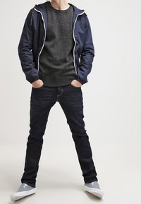 Rompevientos navy con cremallera blanca, suéter de punto gris, jeans azul oscuro y zapatos gris tipo slip-on. Conjunto casual con texturas variadas.