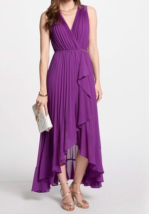 Frau trägt ärmelloses lila plissiertes Wickelkleid mit hohem-niedrigem Saum, goldene Sandalen und hält eine kleine weiße Clutch.