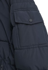 Marinblå pufferjacka ärm med en stor klaffficka och en rund svart knapp. Slät tyg med en kviltad textur.