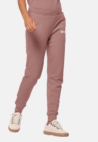 Person wearing dusty pink joggers with Jack Wolfskin logo and off-white lace-up shoes with brown speckled soles, standing on light background.