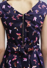 Marindress med färgglada fjärilstryck, dragkedja i ryggen och en rosett knuten i midjan över bar hud på övre ryggen.