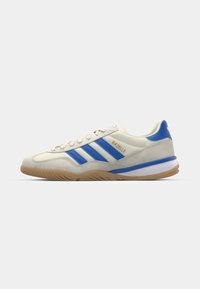 Sneaker met een romige suede bovenkant, blauwe accenten en drie blauwe strepen. Heeft een gumrubberen zool en gouden "Gazelle" branding aan de zijkant.
