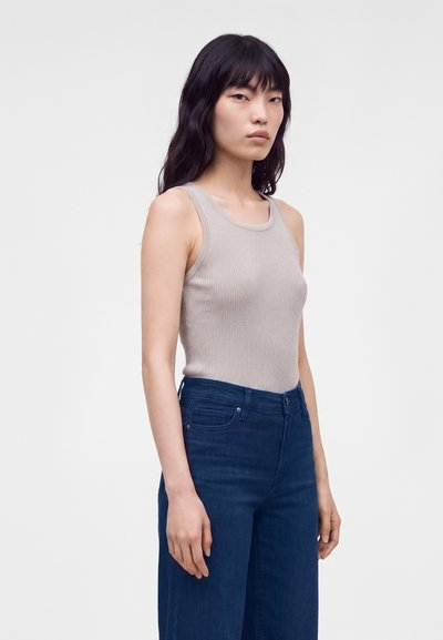 Femme aux longs cheveux noirs portant un haut côtelé beige sans manches et un jean bleu foncé taille haute à jambes larges, debout devant un fond uni.