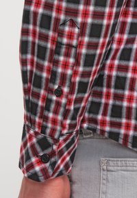 Punainen, musta ja valkoinen ruudullinen pitkähihaisen paidan hiha mustilla napeilla, jonka päällä on vaaleanharmaat farkut.