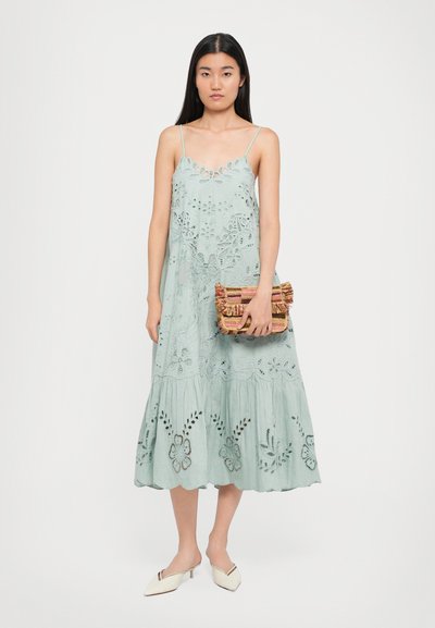 Minttu vihreä brodeerattu mekko ohuilla olkaimilla, kukkakuviot, levenevä helma. Malli pitää käsissään värikästä kudottua clutch-laukkua ja käyttää valkoisia mules-kenkiä.