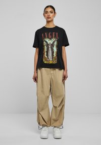Sort bomulds t-shirt med "ANGEL" tekst og grafisk design i lyse farver; parret med løse beige bukser og hvide sneakers.